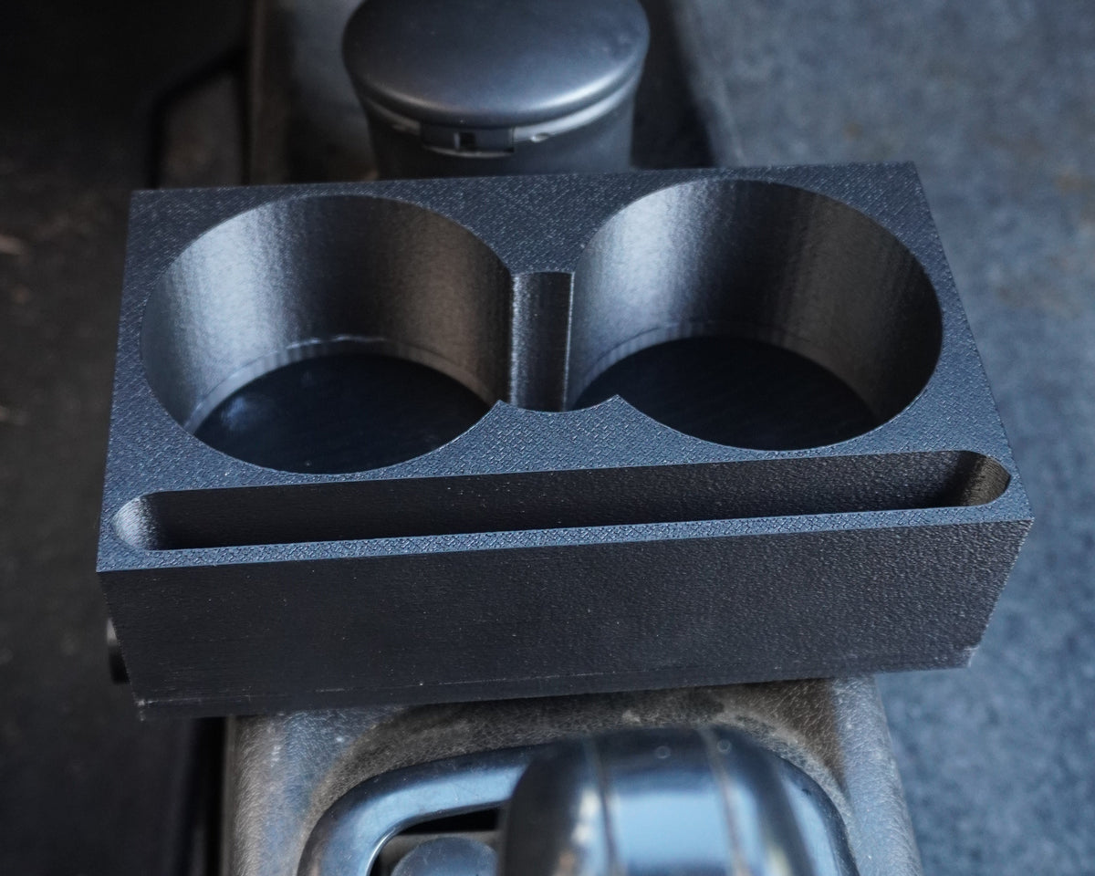 A black dual cup holder tray with a small storage slot, mounted on a car center console next to the gear shift selector.