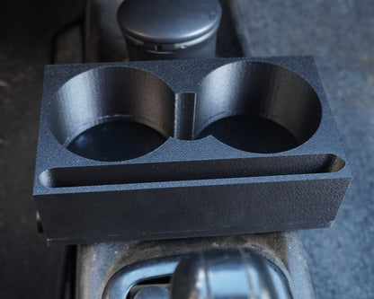 A black dual cup holder tray with a small storage slot, mounted on a car center console next to the gear shift selector.