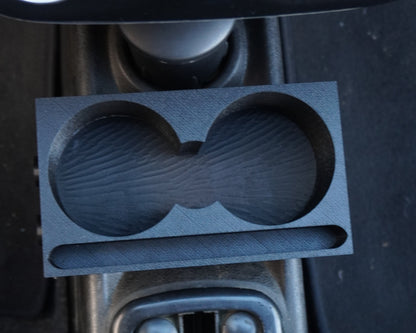 A dark gray dual cup holder insert with a wood-grain texture pattern, shown installed in a car's center console next to the gear shift selector and climate control knobs.