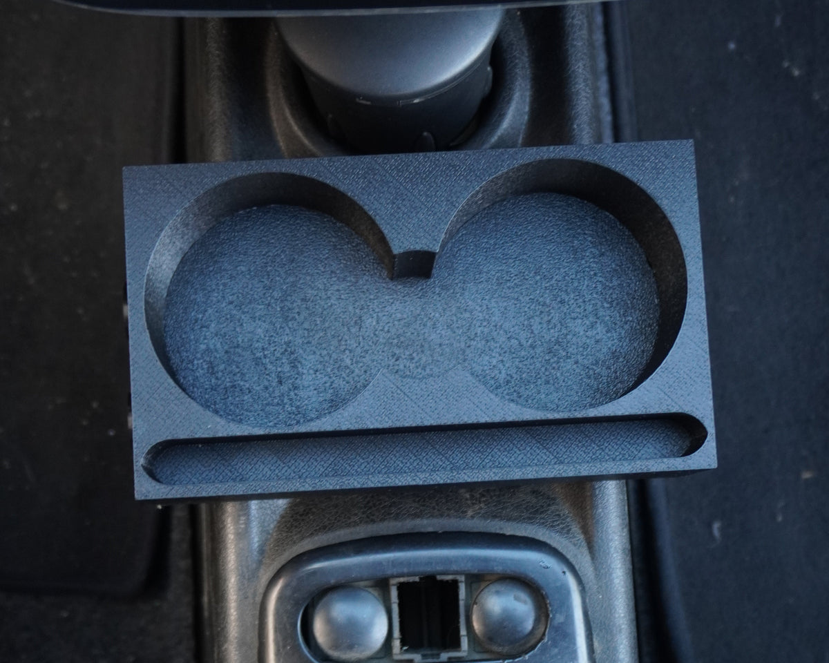 A dark gray double cup holder insert with a textured foam-like surface, shown installed in a car's center console next to the gear shift selector and climate controls.