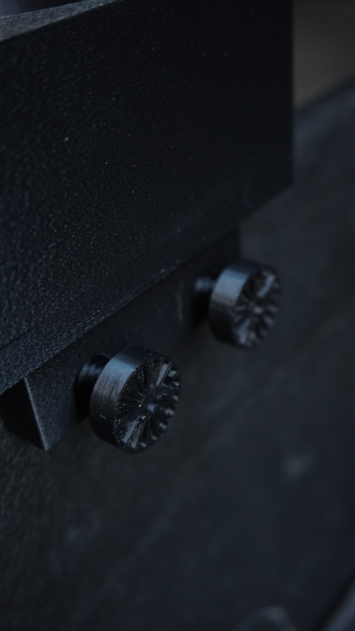 Close-up of a black textured metal surface with two dark metallic knobs or screws, photographed in low light.