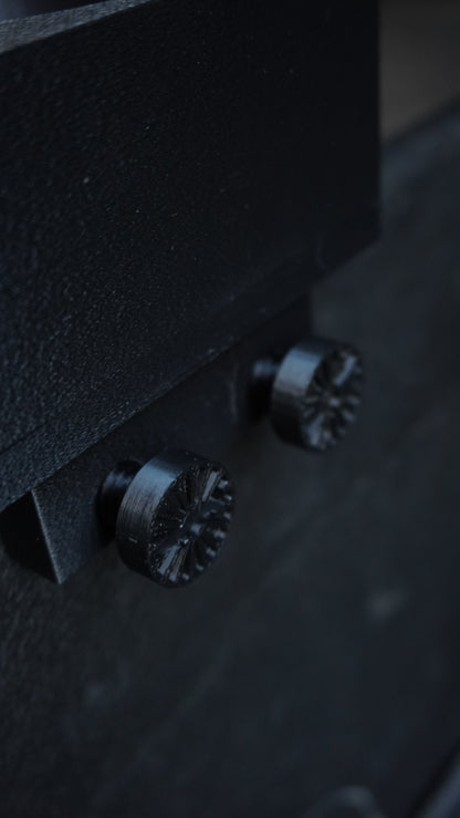 Close-up of a black textured metal surface with two dark metallic knobs or screws, photographed in low light.